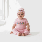 Baby wearing pink Buckhaus Mini Trendsetter short sleeve one piece and matching hat, sitting and smiling against a bright background
